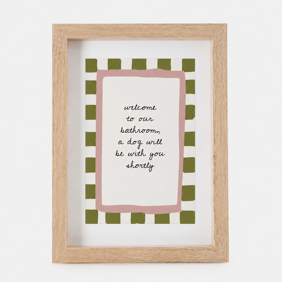 A framed art print with a light wood frame, featuring a pink border and a green-and-white checkered pattern around the text 'welcome to our bathroom, a dog will be with you shortly'.