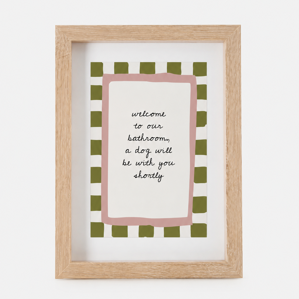 A framed art print with a light wood frame, featuring a pink border and a green-and-white checkered pattern around the text 'welcome to our bathroom, a dog will be with you shortly'.