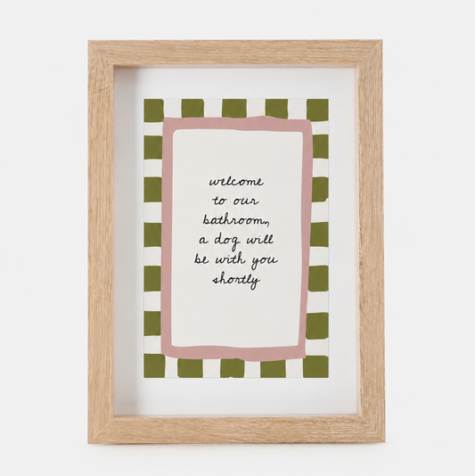 A framed art print with a light wood frame, featuring a pink border and a green-and-white checkered pattern around the text 'welcome to our bathroom, a dog will be with you shortly'.