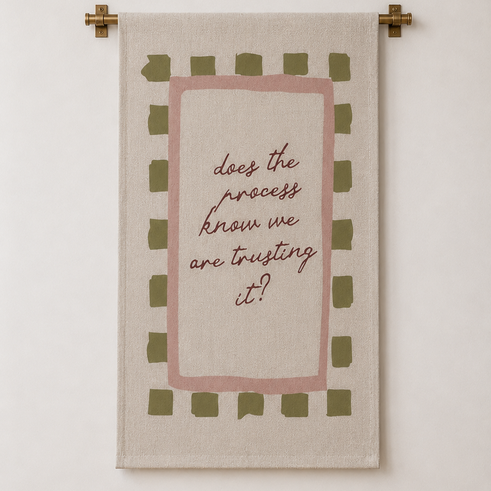 A rectangular wall tapestry with a beige linen-like background, featuring a dusty pink inner border, olive green checkered accents, and the handwritten quote 'does the process know we are trusting it?' in brown script. The tapestry is hung on a cream-colored wall with a light wood floor visible at the bottom.