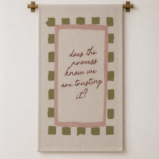 A rectangular wall tapestry with a beige linen-like background, featuring a dusty pink inner border, olive green checkered accents, and the handwritten quote 'does the process know we are trusting it?' in brown script. The tapestry is hung on a cream-colored wall with a light wood floor visible at the bottom.