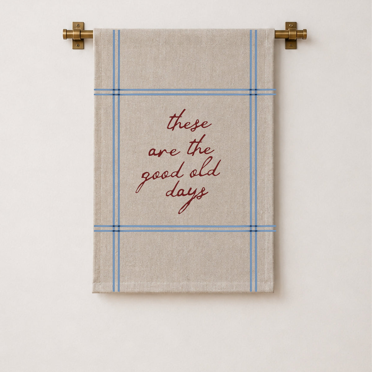 A beige wall hanging with a blue plaid border and the phrase 'these are the good old days' in red script, hanging from a brass rod against a white wall.