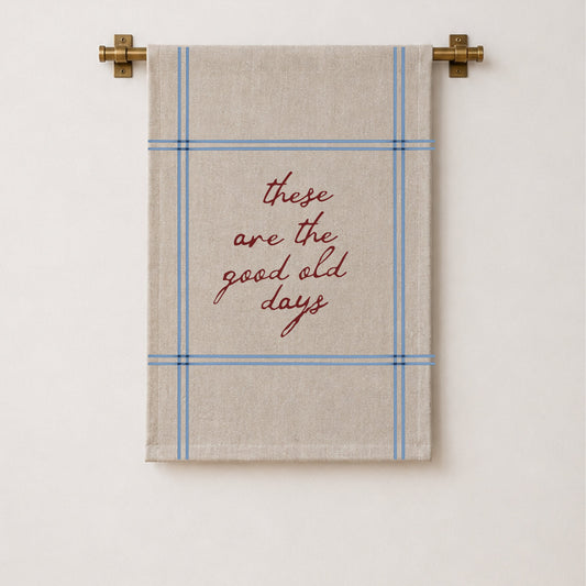 A beige wall hanging with a blue plaid border and the phrase 'these are the good old days' in red script, hanging from a brass rod against a white wall.