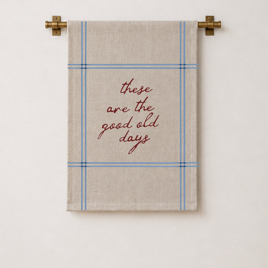 A beige wall hanging with a blue plaid border and the phrase 'these are the good old days' in red script, hanging from a brass rod against a white wall.