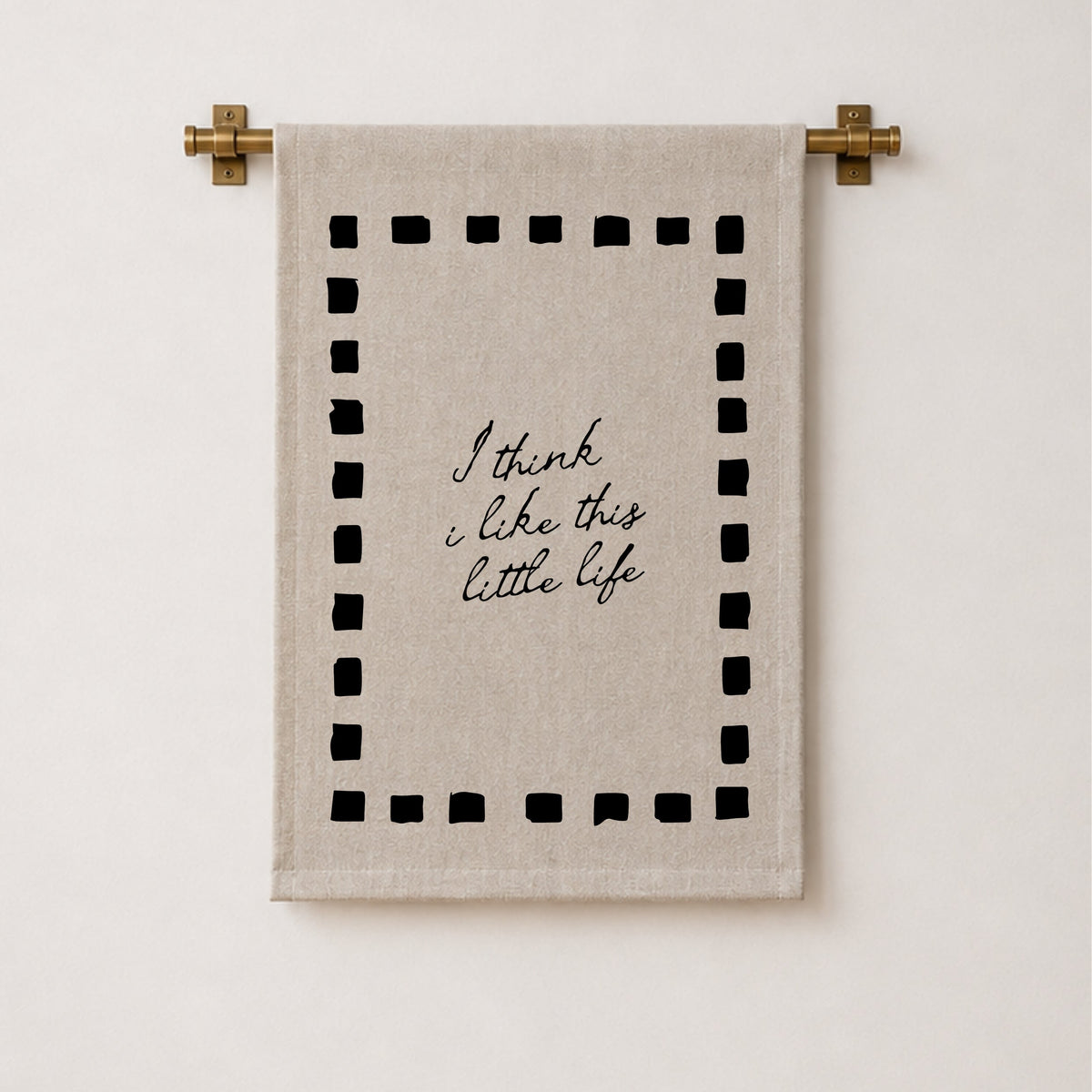 A beige linen tea towel with black embroidered text reading 'I think i like this little life' inside a dotted border, hanging from a brass rod against a white wall.