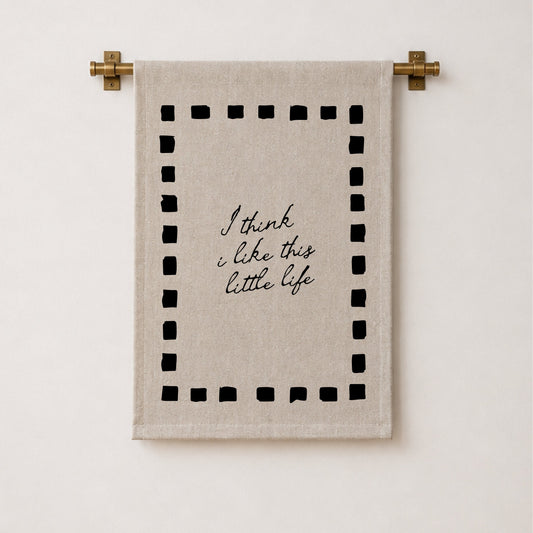 A beige linen tea towel with black embroidered text reading 'I think i like this little life' inside a dotted border, hanging from a brass rod against a white wall.